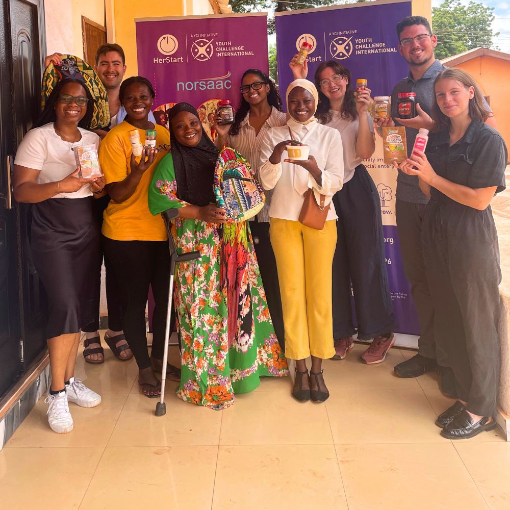 A group of young women and men, including young women entrepreneurs from Tanzania and Canadian volunteers, standing on a tiled floor and holding up products like handmade backpacks, food products, and spices, all while smiling at the camera.