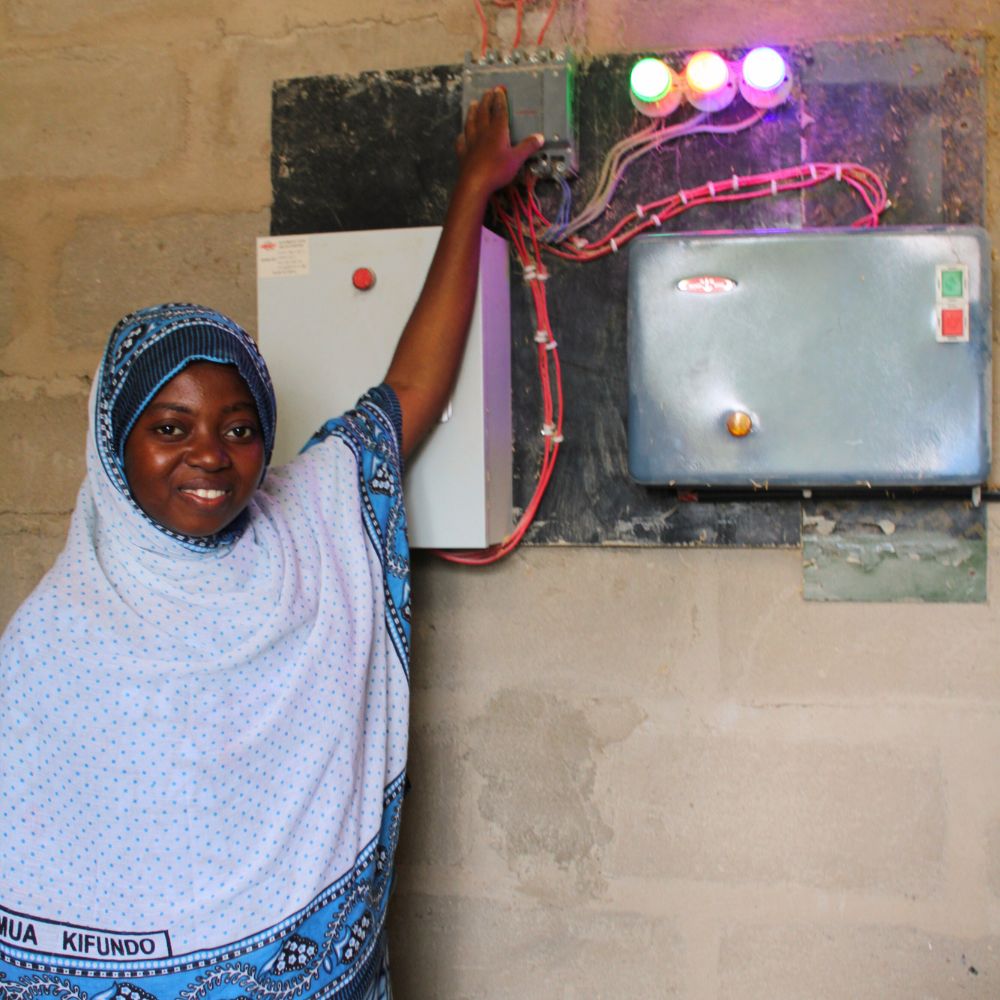 1000x1000 (7) A woman in a blue and white hijab stands by an electrical control panel with colorful indicator lights.