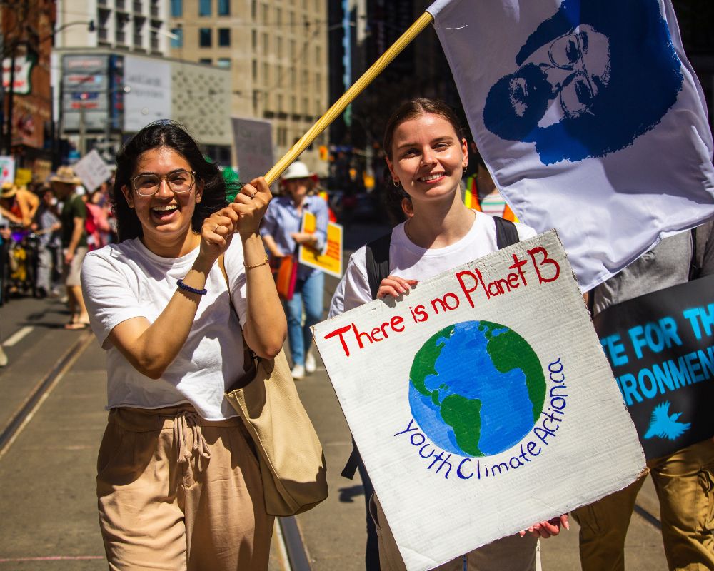 1000x800 (4) Two smiling young women at a climate protest hold a “There is no Planet B” sign and a flag with a face on it.