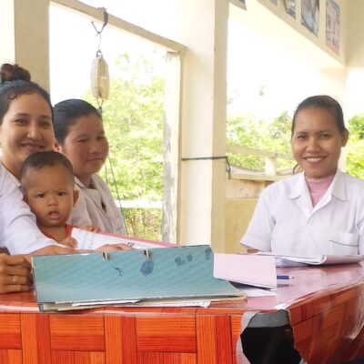 Six adults and a baby sit at a red table in a bright room, smiling at the camera.