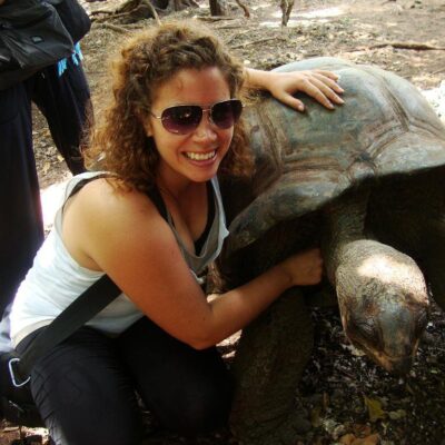 Woman smiling and hugging a large tortoise outdoors, with other people standing in the background.