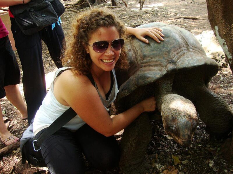 22-Giant-Tortoise Woman smiling and hugging a large tortoise outdoors, with other people standing in the background.