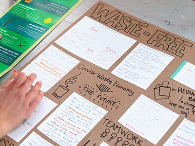 A person arranges notes and cards on a table, with a sign reading "WASTE-FREE TORONTO" visible.