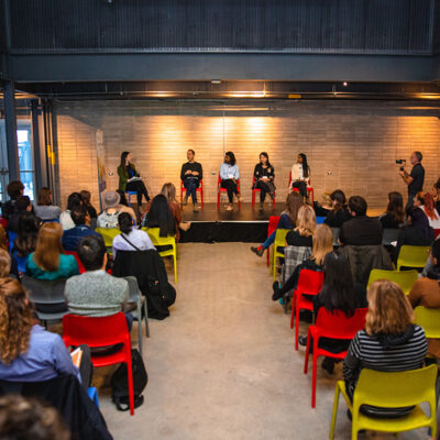 A panel of five people speaks to an audience in a modern indoor event space with colorful chairs.