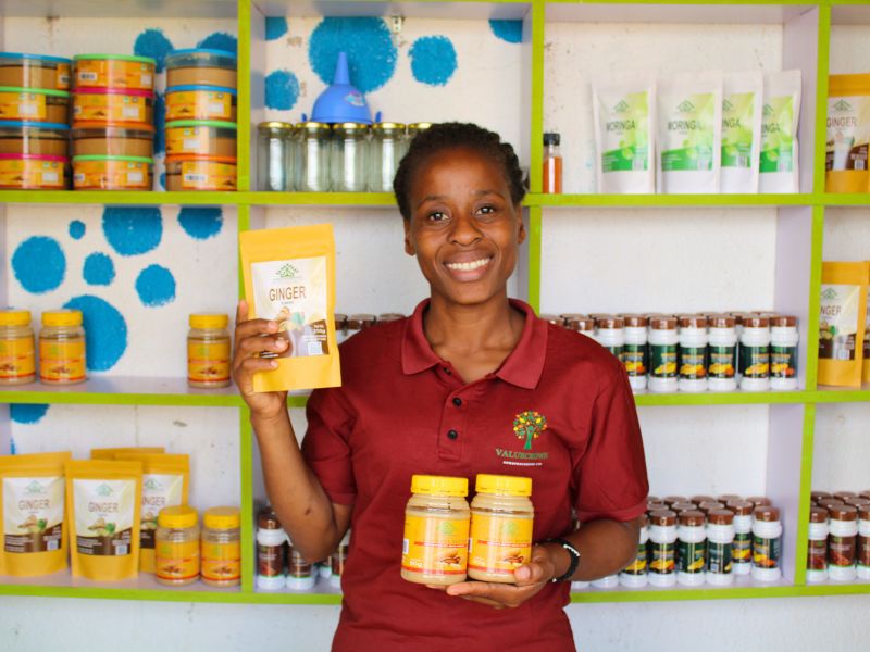 800x600 (4) A woman in a red shirt smiles, holding jars and a pack of ginger in a shop with spice products on shelves.