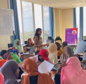 A group of people in colorful attire attend a workshop in a bright room with large windows and a whiteboard.