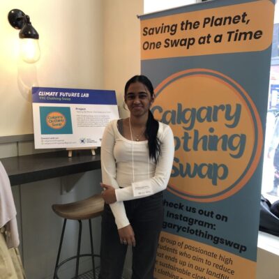 A woman stands smiling beside a "Calgary Clothing Swap" banner at an indoor event.