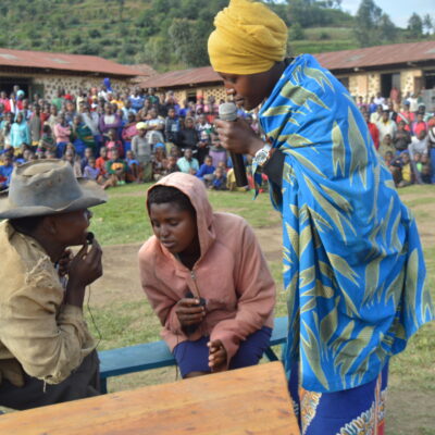 Three people perform in front of a crowd outdoors, with one person holding a microphone and wearing a yellow headscarf.