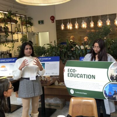 Two women present an eco-education poster to an audience in a modern, plant-filled room.