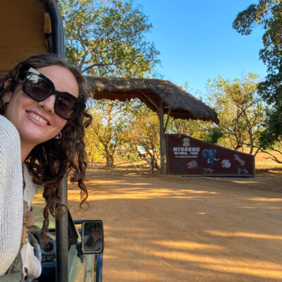 A smiling woman in sunglasses leans from a safari vehicle near the Nyerere National Park entrance sign.