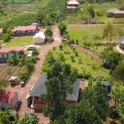 An aerial shot of a village in an African country, including concrete shelters with red and black roofs, red dirt roads and lush green lawns and trees.