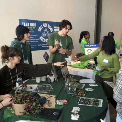 Four people stand at a table with plants and info, discussing eco-friendly biking at an event.