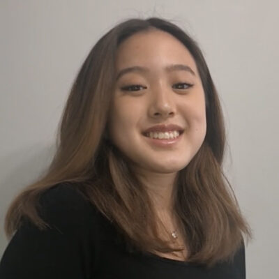 A young woman with long brown hair smiles at the camera against a plain light-colored background.