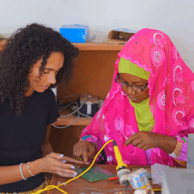 Two young women, one with curly black hair and the other wearing a vibrant pink headscarf, working on connecting an electric circuit board.