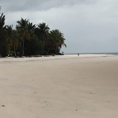 Woman with a backpack smiles while walking along a deserted tropical beach with palm trees and a boat in the distance.