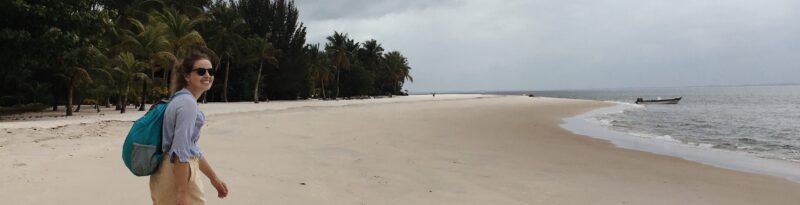 Lauren-blog-header-e1530204840556 Woman with a backpack smiles while walking along a deserted tropical beach with palm trees and a boat in the distance.