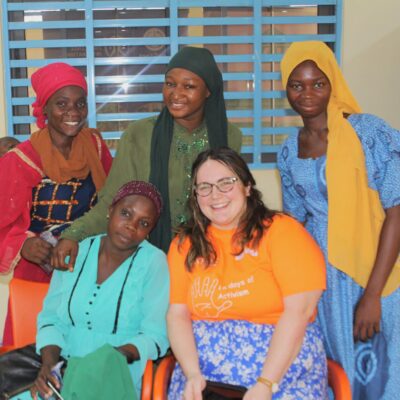 Five women, smiling, pose together indoors; one wears an orange shirt and sits in front of the others.