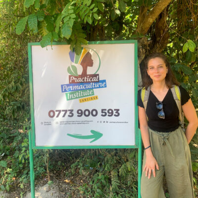 Woman standing next to a Practical Permaculture Institute Zanzibar sign outdoors, surrounded by greenery.