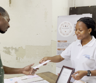 A woman in a white shirt hands a paper to a man across a table with documents and a framed certificate.