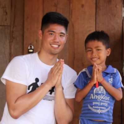 A man and a young boy smile and press their palms together in greeting, standing in a wooden doorway.