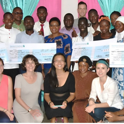 A diverse group poses indoors with three large ceremonial checks and colorful pink and teal curtains behind them.