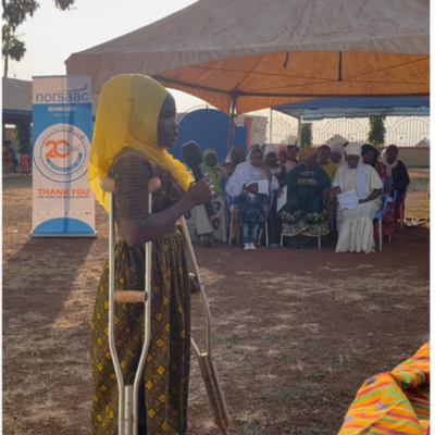A young woman on crutches speaks into a microphone at an outdoor event under a tent.