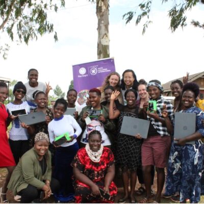 A joyful group of women and girls posing outdoors, some holding laptops and phones, in front of a purple banner.