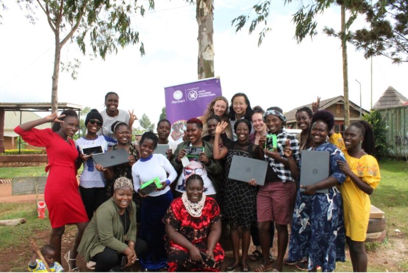 TARA-recipients-and-volunteers-2023 A joyful group of women and girls posing outdoors, some holding laptops and phones, in front of a purple banner.