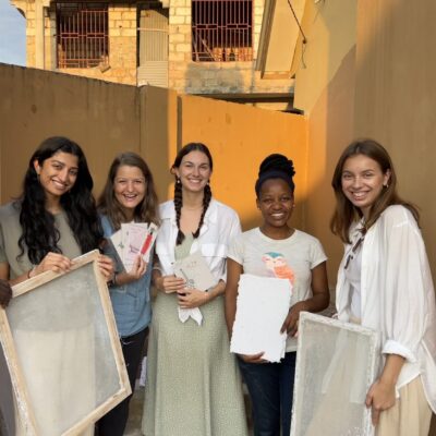 Five women standing against a yellow wall at golden hour, all holding various paper-making materials like frames, paper and cards.