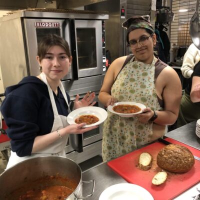 Two people in a kitchen hold bowls of soup, with bread and a pot of soup on the counter in front of them.