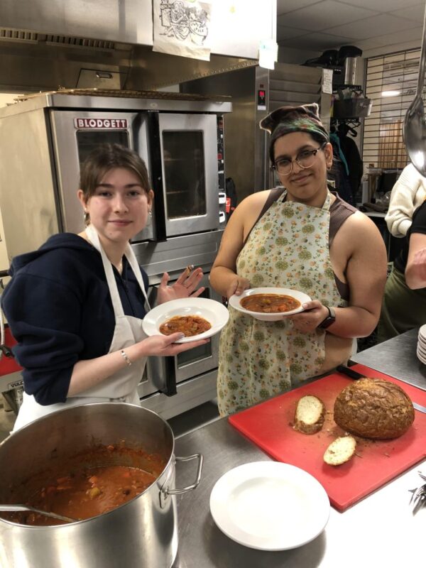 Two people in a kitchen hold bowls of soup, with bread and a pot of soup on the counter in front of them.