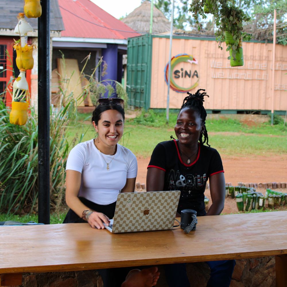 Conception sans titre (6) Deux femmes souriant et travaillant sur un ordinateur portable à une table en plein air avec de la verdure et des bâtiments en arrière-plan.