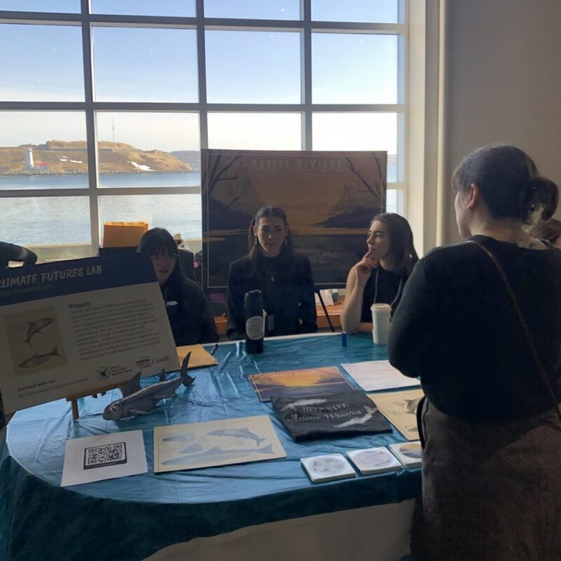 YCI-Innovators_2-min-1024x1024 Four people converse at an information table by a window with an ocean view and display boards.