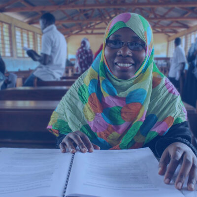 Smiling woman in a colorful hijab reads an open book in a classroom with other students in the background.