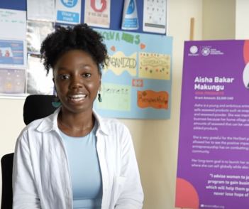 A woman sits and smiles next to a purple informational poster and a wall with colorful signs and papers.