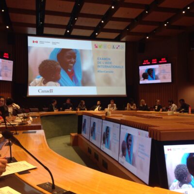 A conference room with people seated, screens display “Examen de l’aide internationale” and a smiling woman and child.