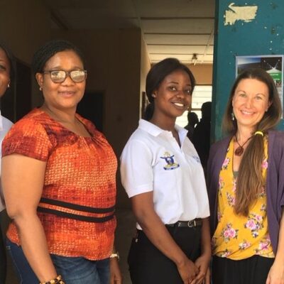 Seven women stand smiling together, six in matching white shirts, in front of a blue door.