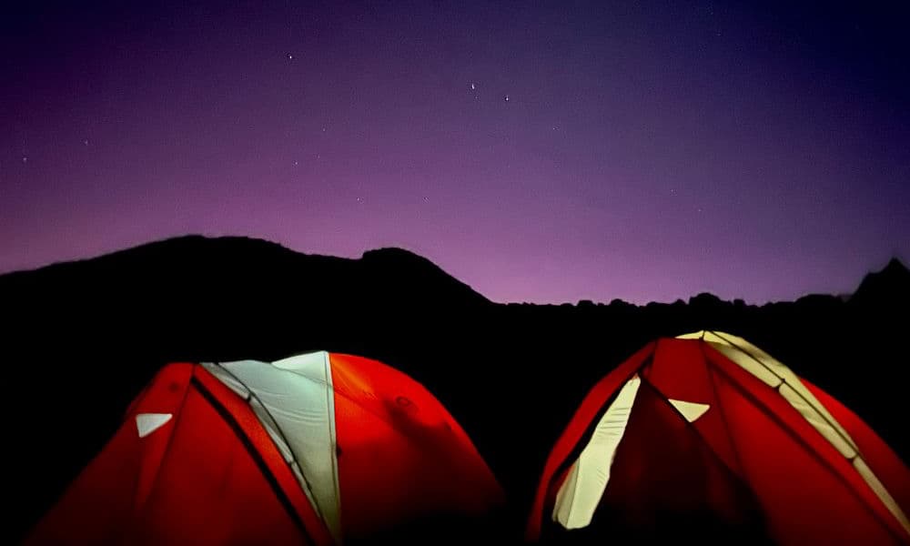 kili-climb-2025-2 Deux tentes rouges brillent dans l'obscurité sous un ciel violet étoilé avec des collines en silhouette à l'arrière-plan.