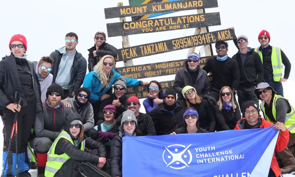 kili-climb-2025-3 Un groupe pose au sommet du Kilimandjaro, souriant et tenant une bannière de Youth Challenge International.