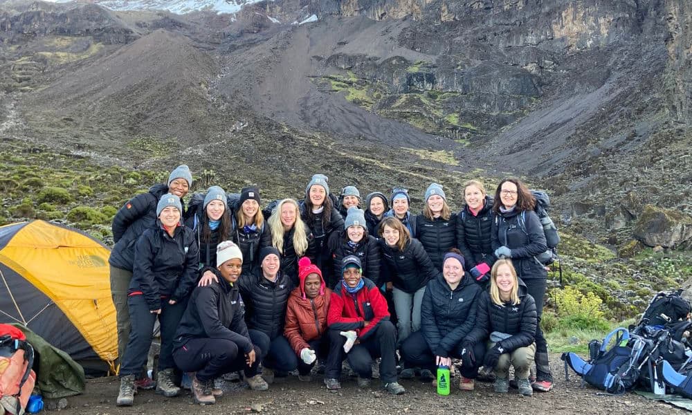 kili-climb-2025-4 Un groupe de randonneurs souriants, vêtus d'équipements de plein air, posent ensemble près d'une tente au pied d'une montagne rocheuse.