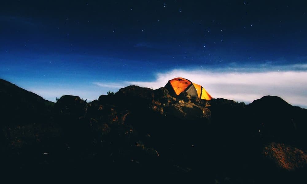 kili-climb-2025-5 Tente orange sur un terrain rocailleux, la nuit, sous un ciel étoilé avec quelques nuages en arrière-plan.