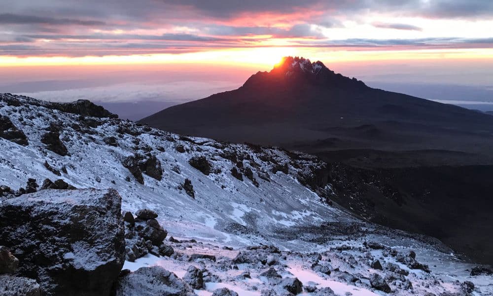 kili-climb-2025-6 Pente de montagne enneigée avec terrain rocheux au lever du soleil, le soleil se levant derrière un sommet lointain sous un ciel nuageux.